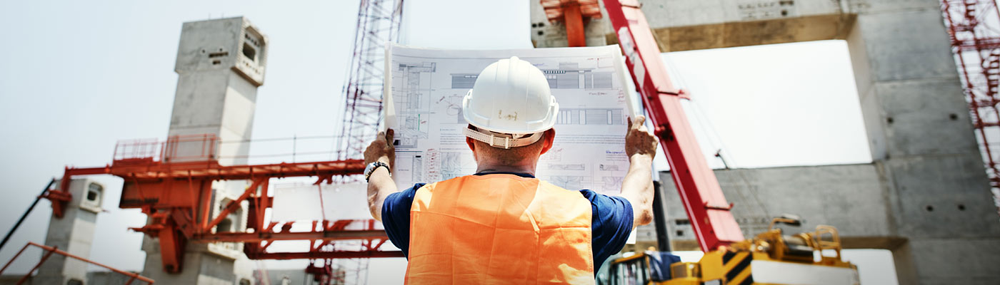 Ein Bauarbeiter hält einen Bauplan in einer Baustelle mit Kränen und Betonstrukturen im Hintergrund.